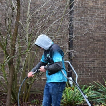 Prime & Shine technician adjusting pressure washer connections in a garden bed beside a brick building