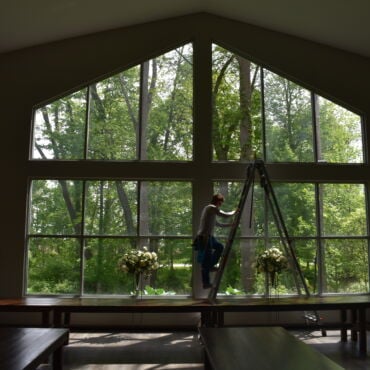 Technician on a ladder cleaning the tall cathedral-style windows of a banquet hall with a wooded view and floral arrangements below