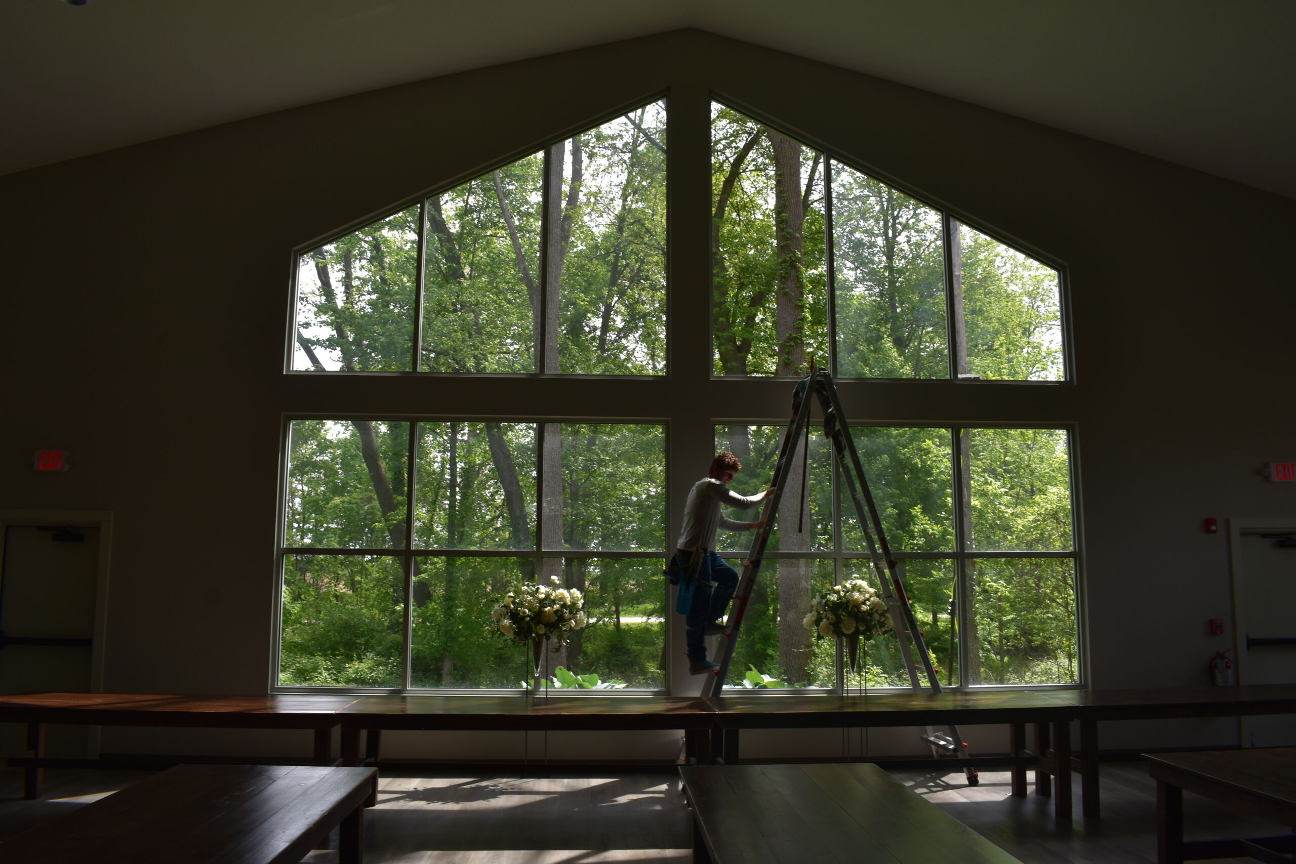 Technician on a ladder cleaning the tall cathedral-style windows of a banquet hall with a wooded view and floral arrangements below