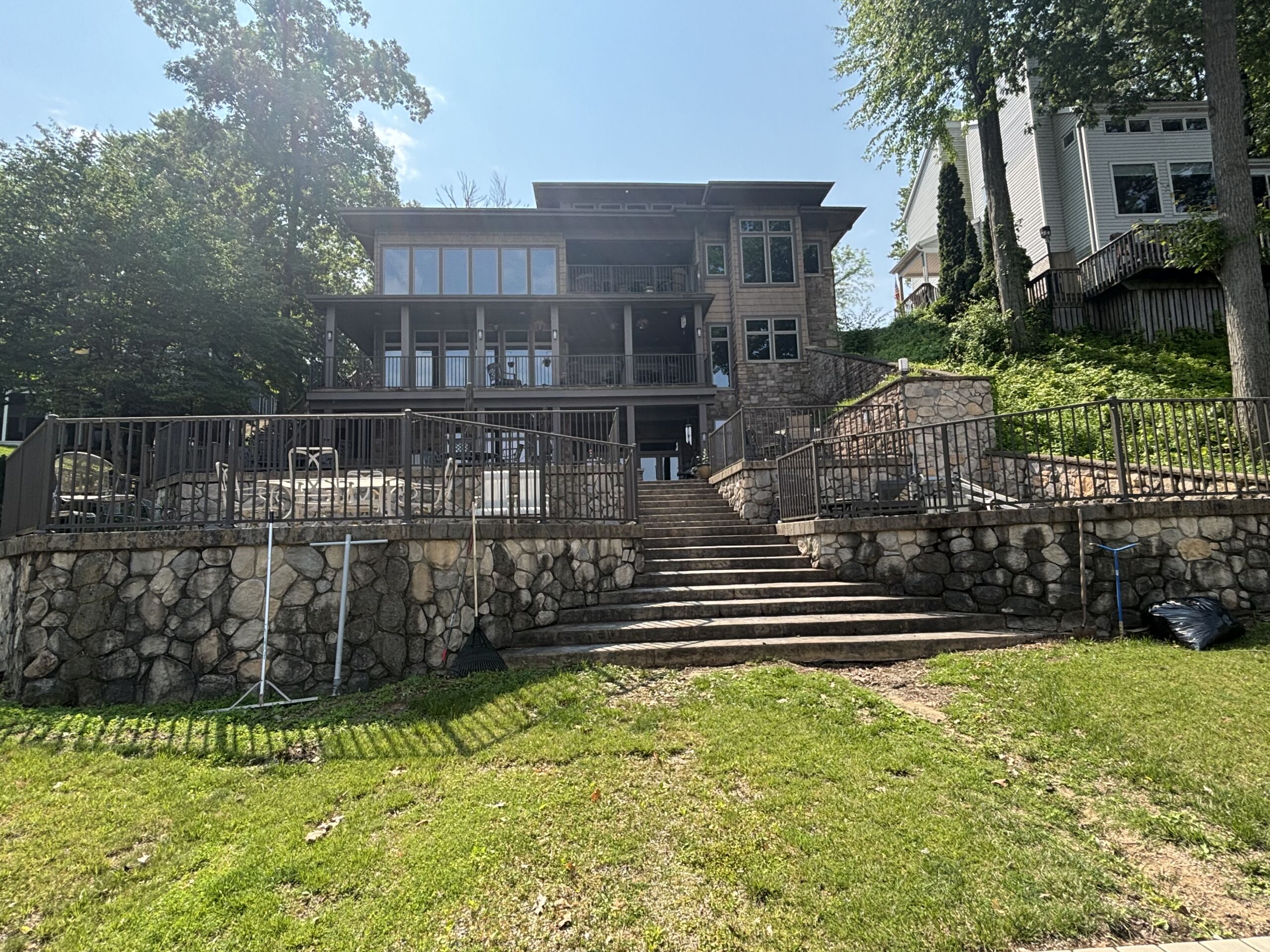 Rear view of a large lakefront home with multi-level stone retaining walls, wide concrete steps, and wraparound decks ready for exterior cleaning