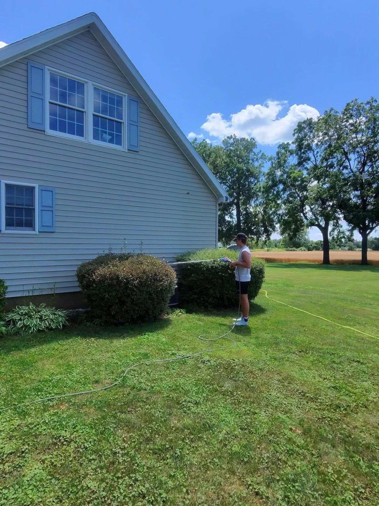 Technician soft washing the vinyl siding of a two-story farmhouse surrounded by open fields on a sunny summer day