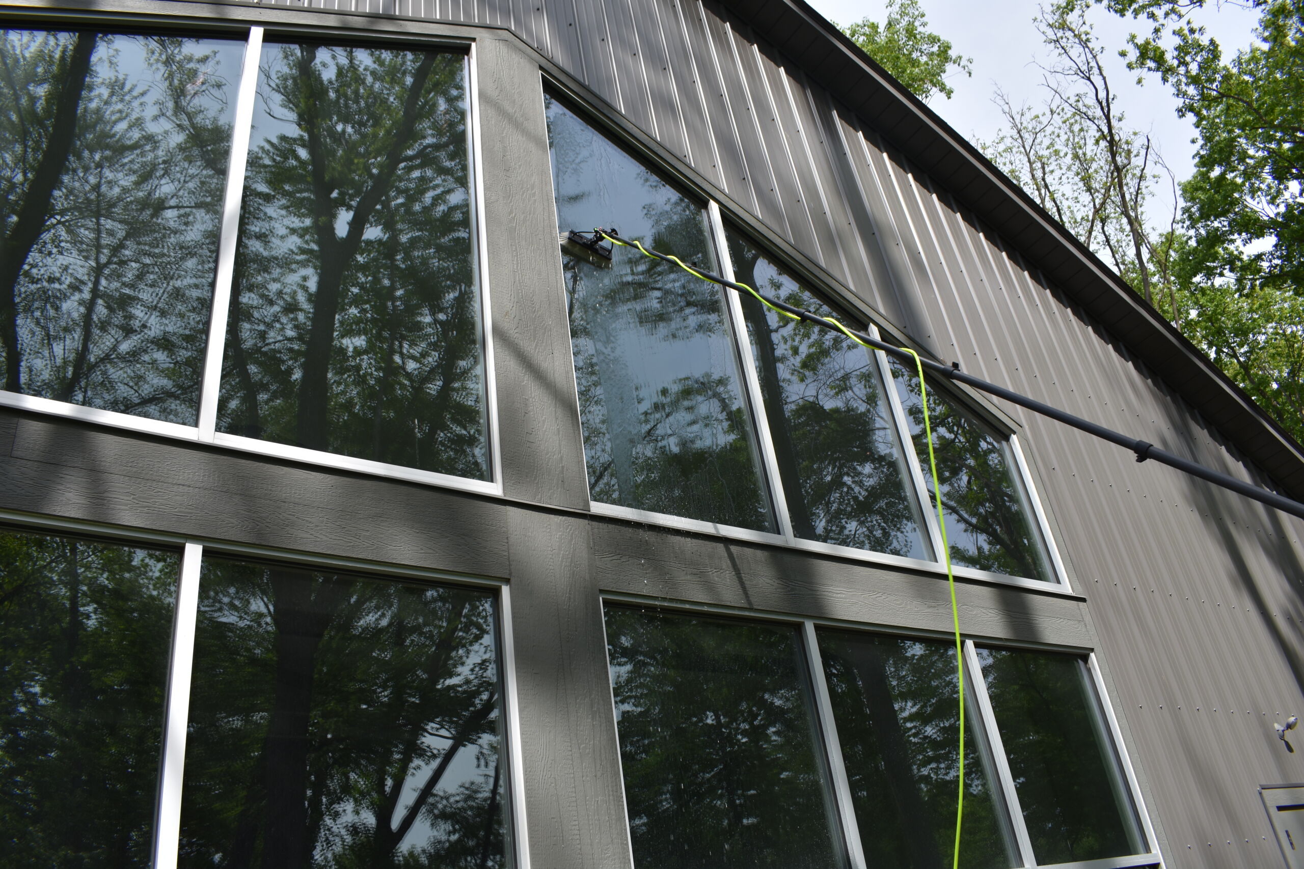Technician using an extended water-fed pole to clean large floor-to-ceiling windows on a modern dark-sided building