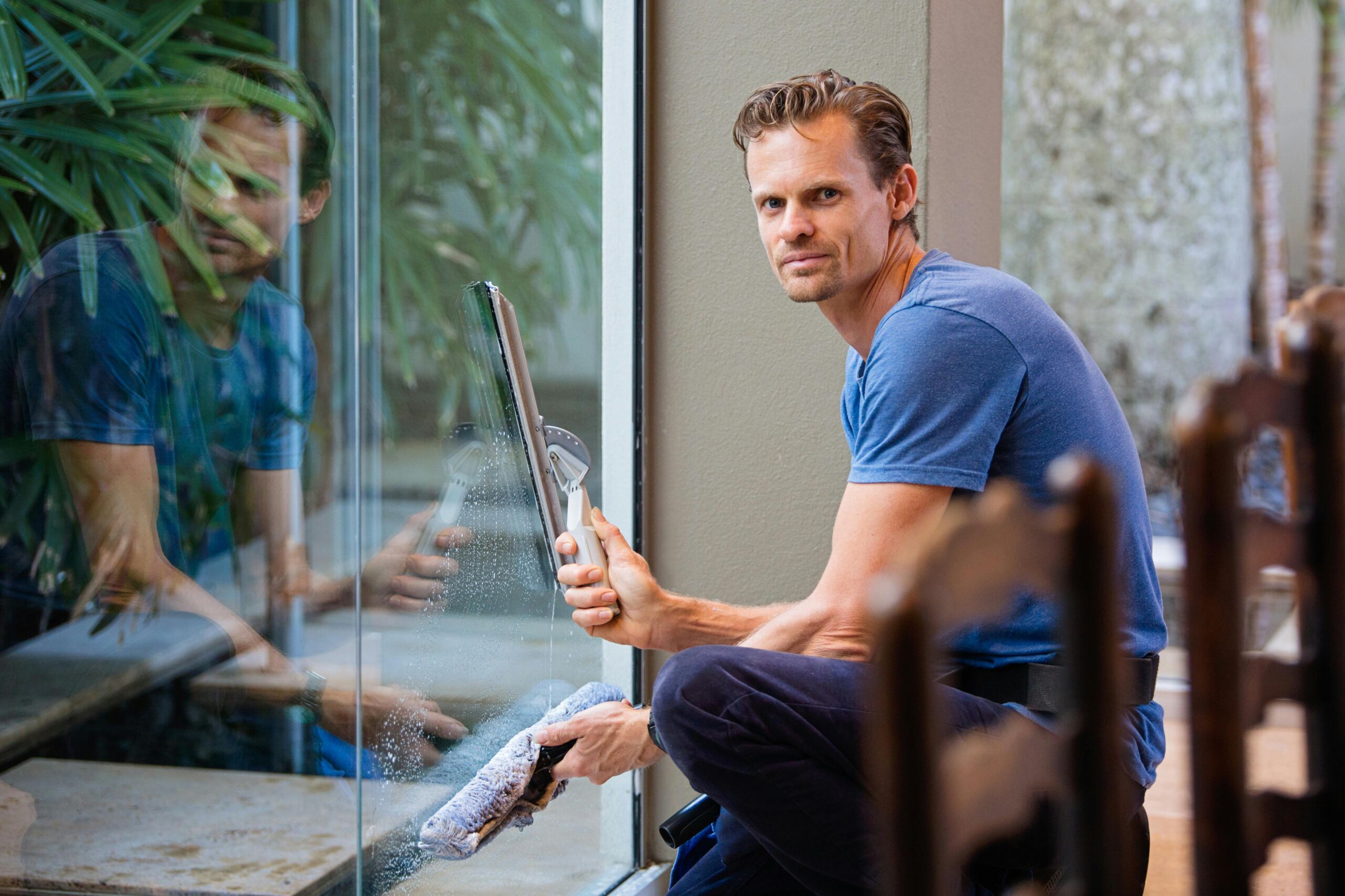 Window cleaner using a squeegee and scrubber to clean a large glass pane with his reflection visible on the surface