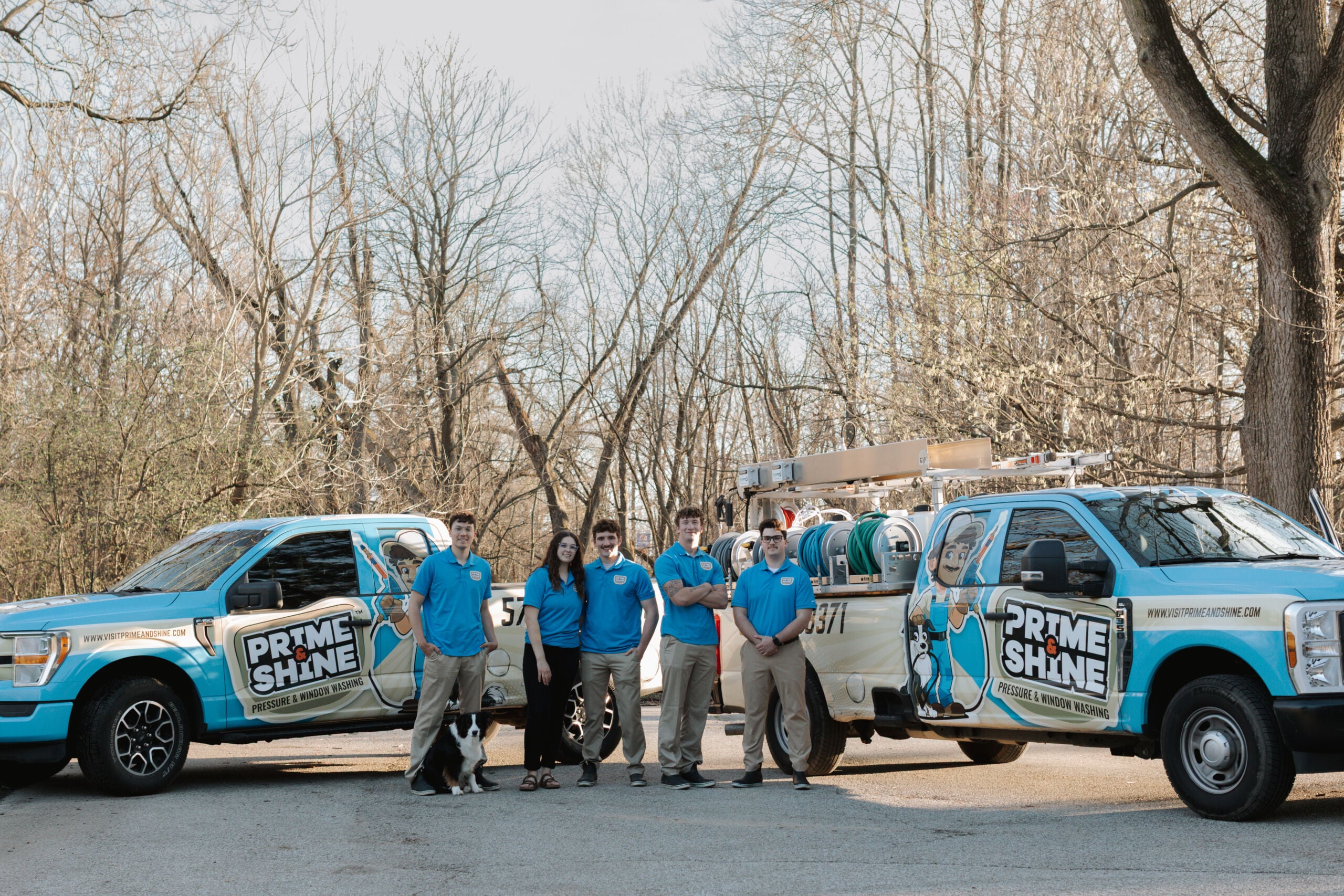 The Prime & Shine team in blue uniforms posing together with their border collie between two branded service trucks