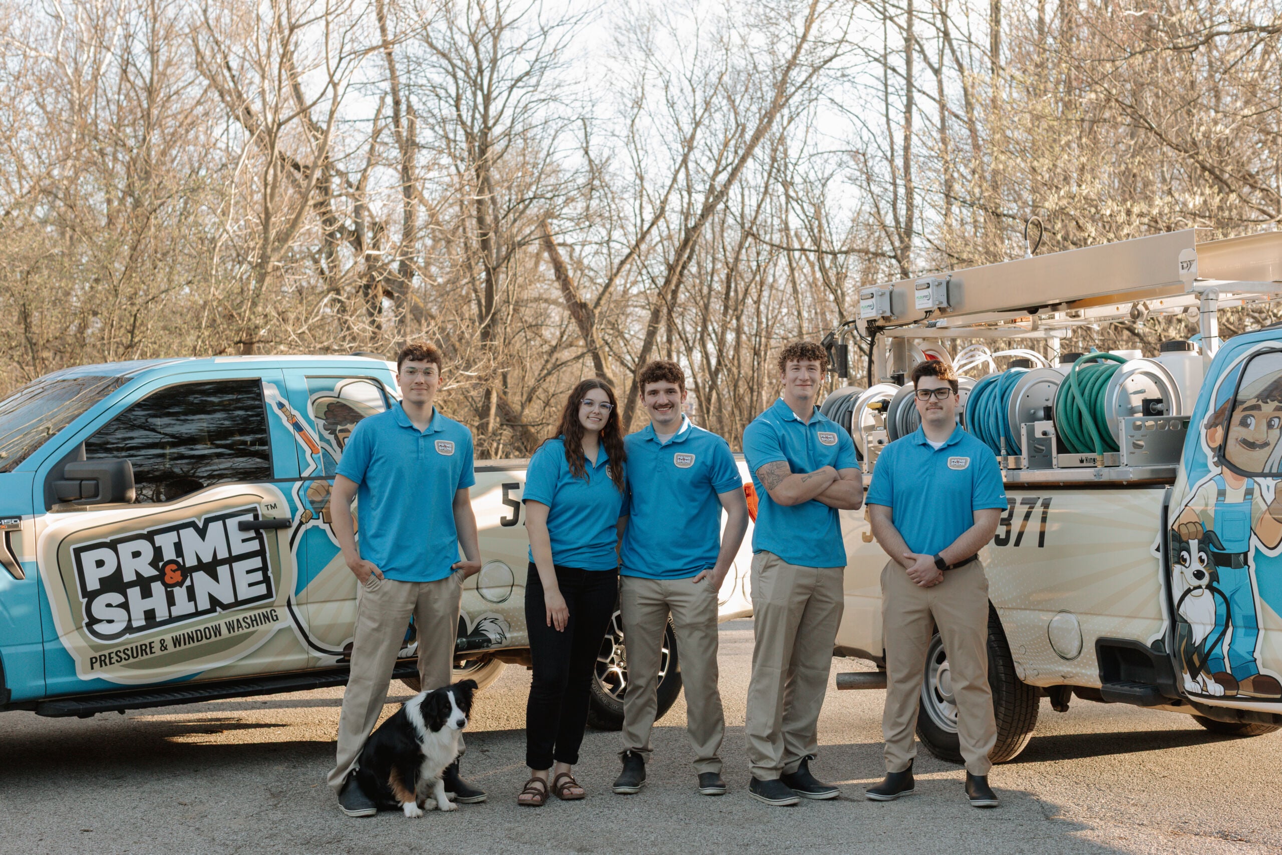 Full Prime & Shine crew in matching blue uniforms standing together with their border collie mascot between two fully equipped branded service vehicles