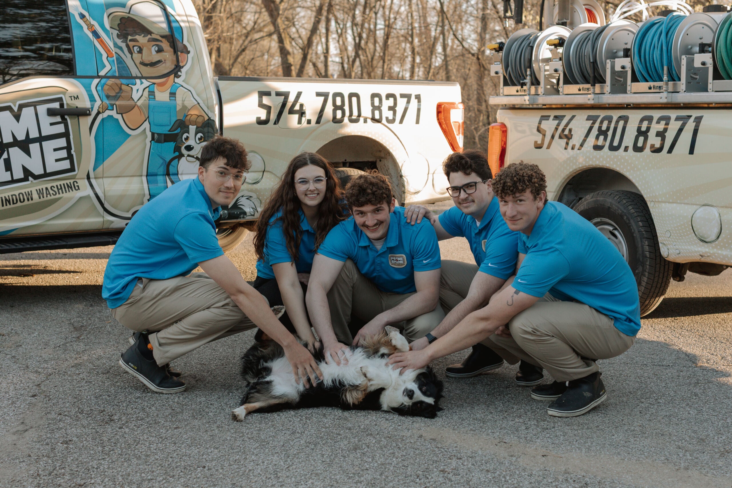 Prime & Shine crew laughing and petting their border collie belly-up in front of two branded service trucks