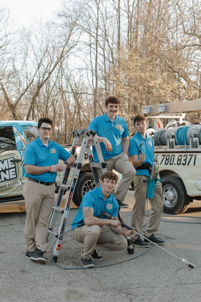Four Prime & Shine male crew members posing confidently with a ladder, squeegee, and pressure wand in front of branded service trucks
