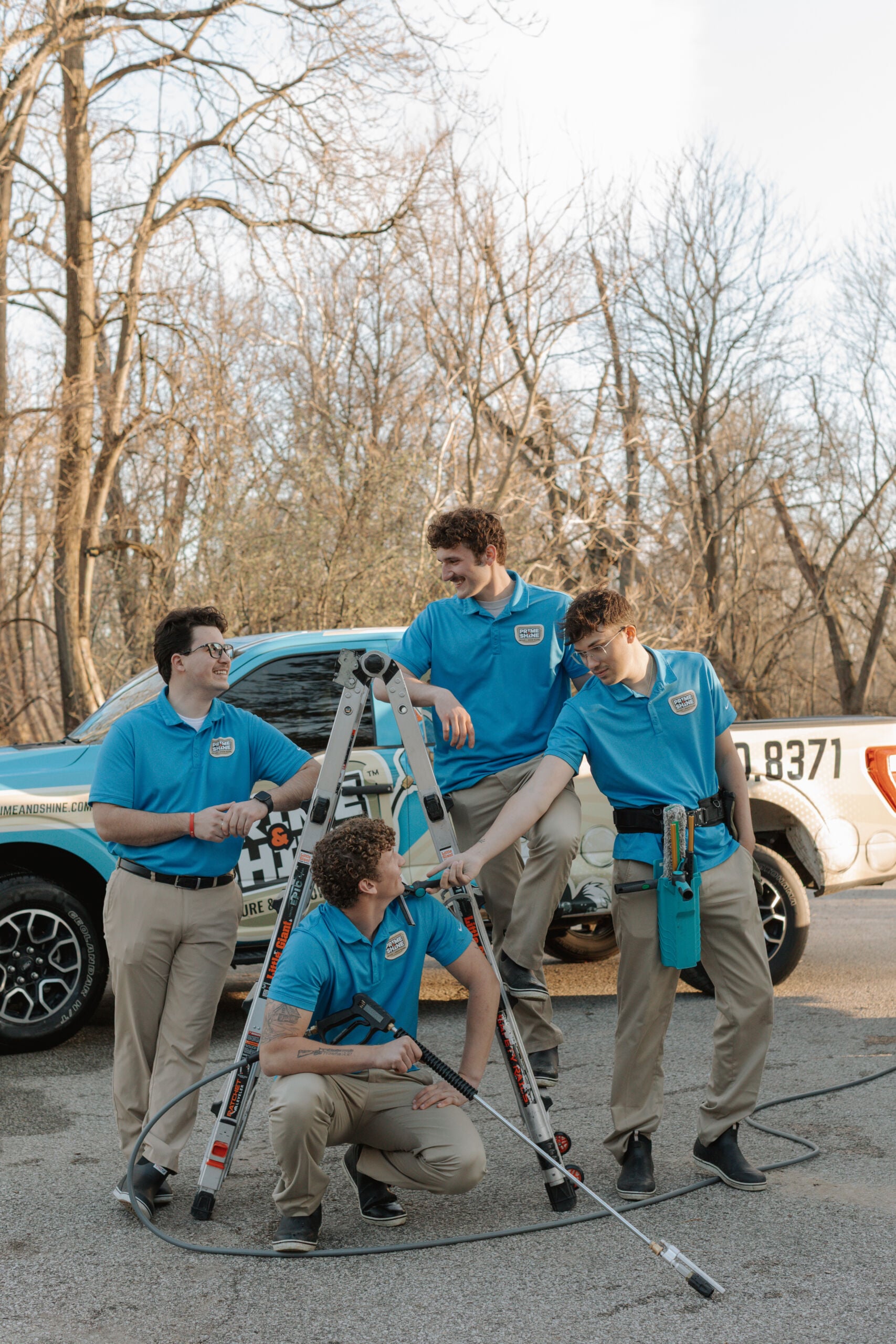 Four Prime & Shine crew members laughing together around a ladder and pressure wand in front of their branded truck
